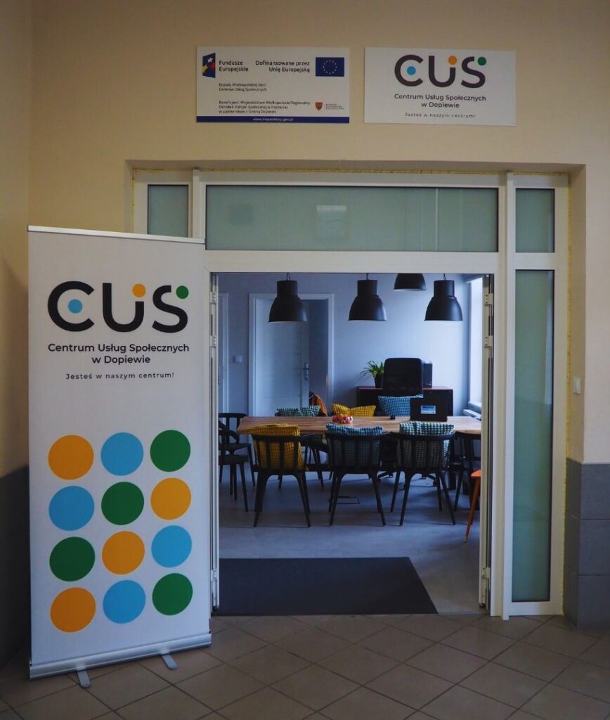 Zdjęcie przedstawia wejście do części administrowanej przez Centrum Usług Społecznych w Dopiewie. Obok drzwi stoi baner z logo CUS.